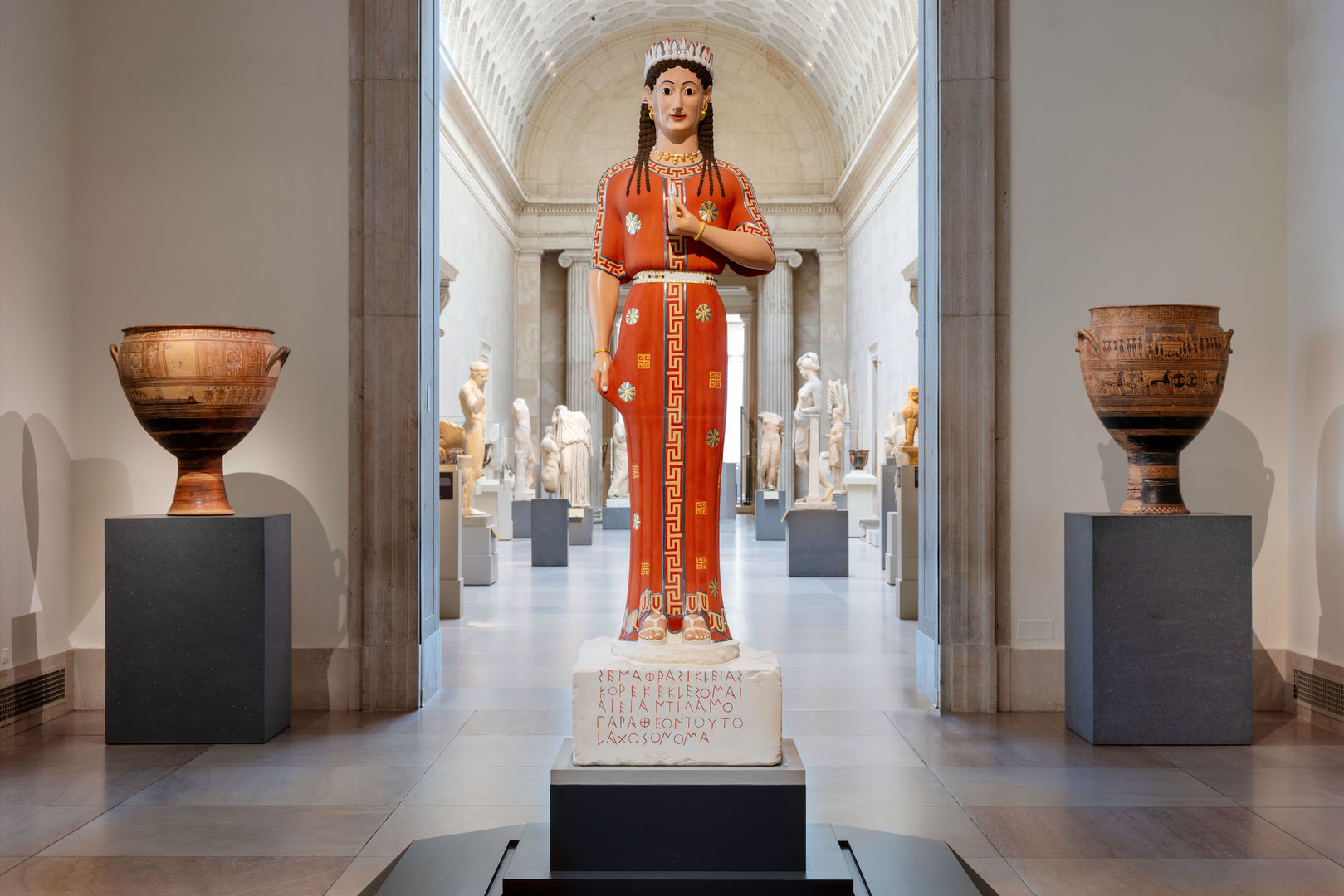A brightly-painted statue of a woman stands atop a pedestal inscribed with Greek letters. In the background sit two ornate clay urns and many white marble statues.